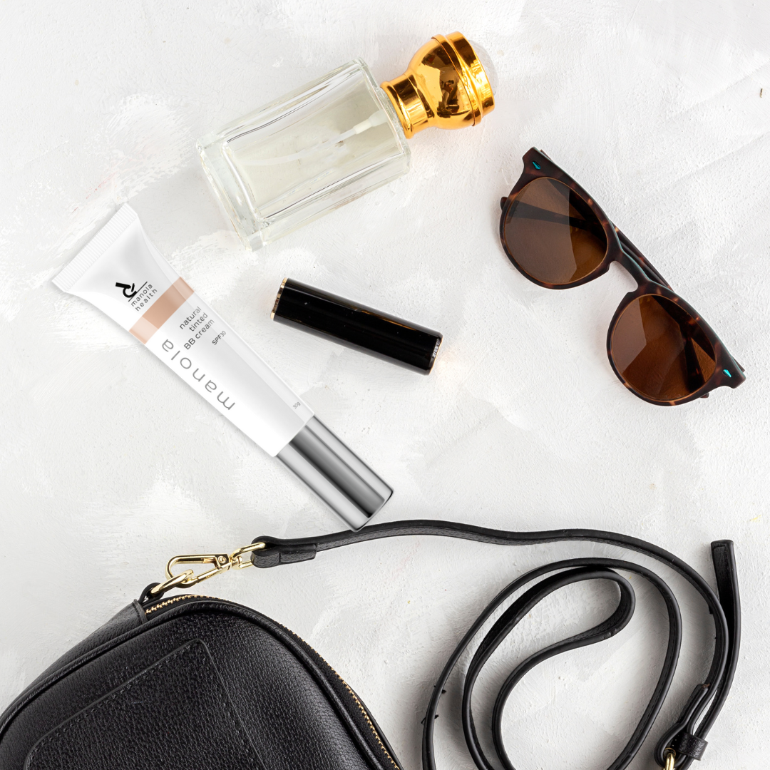 A styled flat-lay of various fashion accessories and beauty items on a textured white background, including a black pebbled leather crossbody bag, tortoiseshell sunglasses, a perfume bottle with a gold cap, a tube of Momo La makeup, and a black cylindrical case.