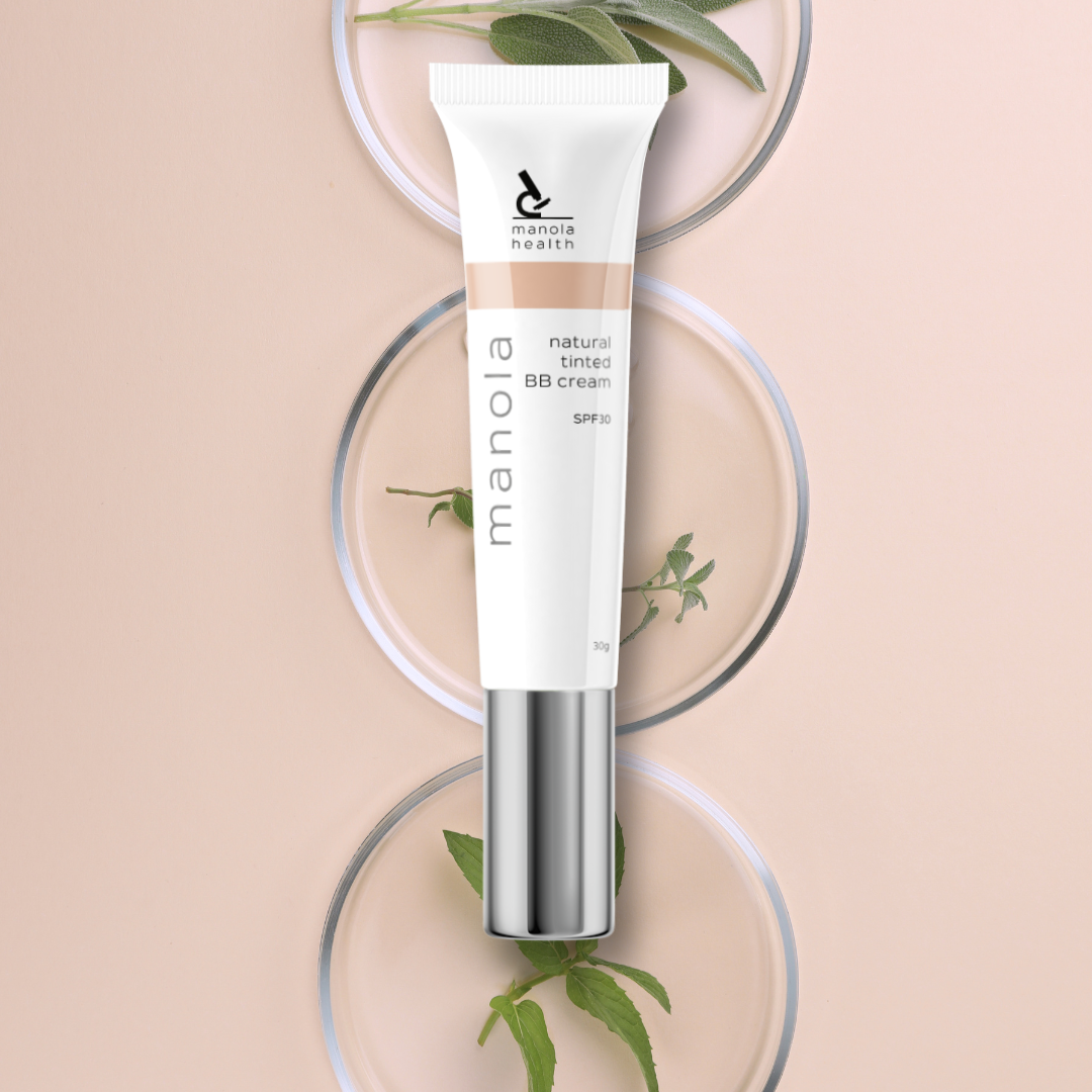 A squeeze tube of Manola BB cream in a natural tint shade, displayed vertically on a light peach background with decorative glass dishes and green leaves.