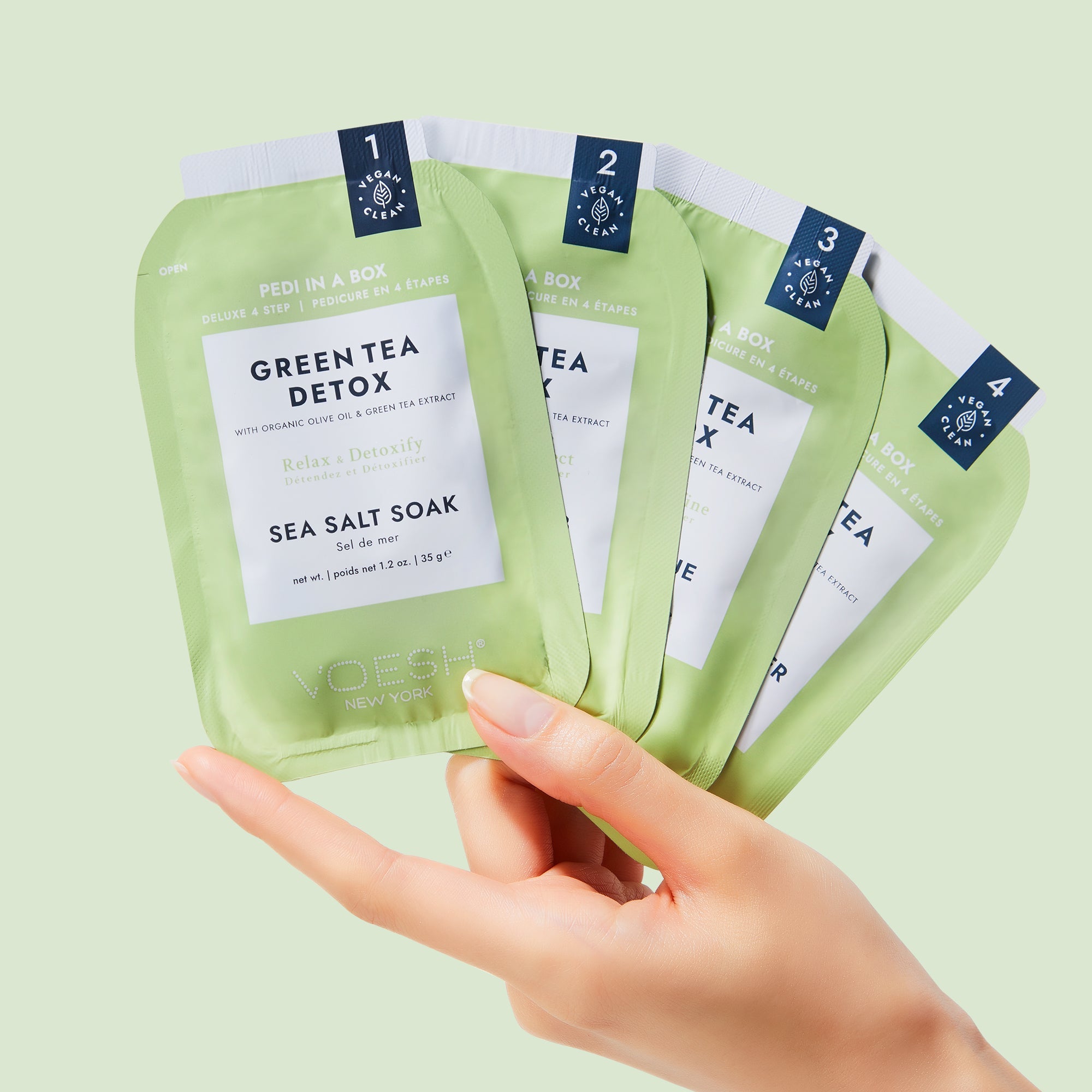 Hand holding green tea detox sea salt soak packages against a light green background