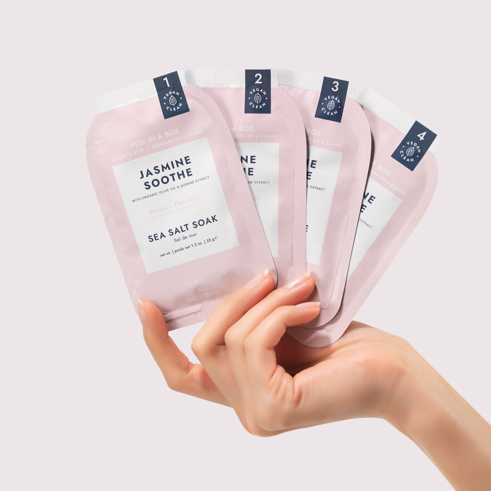 Hand holding four pink 'Jasmine Soothe' sea salt soak packages against a light background